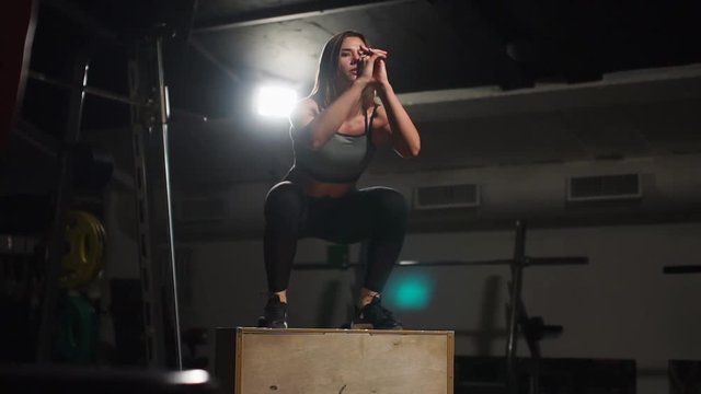 Beautiful female fitness athlete performs box jumps in a dark gym wearing black sports top and short tights with face hidden