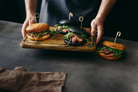 Man holding on cutting board set black burger and burger with classic bun with beef medium rare, spinach, green sauce and micro greens