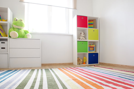 Colourful children rooom with white walls and furniture. Rainbow carpet at home interior with a window.