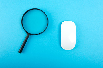 Magnifying glass and white pc mouse on blue table. Top view.