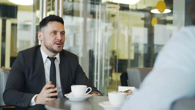 Cheery Caucasian businessman gesticulating and rejoicing in buisness profits while talking to his partner in mod cafe during lunch.