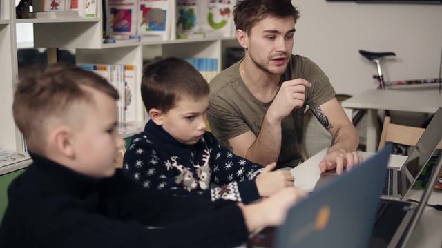 Attarctive male programmer teacher with several tattooes on his arms explaining something to his two little pupils boys on a programming class. Alternative education for children.