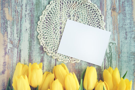 bouquet of fresh yellow tulips on a multi-colored wooden background with a blank card for text on a lacy round napkin