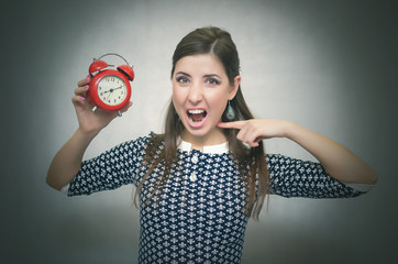 Angry woman holding in hands a red alarm clock and showing on it by her finger and shouting. Wicked boss or teacher. Be late at work or school. Oversleep concept.