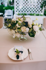 Beautiful and exquisite decoration of the wedding celebration in in the middle of a green garden. Banquet served table with a beige tablecloth, plates, napkins, cards and flower compositions