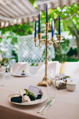 Beautiful and exquisite decoration of the wedding celebration in in the middle of a green garden. Banquet served table with a beige tablecloth, plates and candlesticks with blue candles under cover