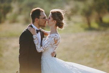 Beautiful wedding photosession. Handsome groom in a black suit and young bride in white lace dress with exquisite hairstyle on walk around the big green field against the trees and bushes background