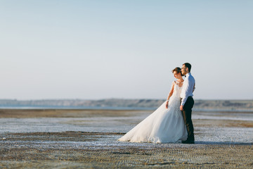 Fototapeta na wymiar Beautiful wedding photosession. Handsome unshaved groom in a black trousers and young cute bride in white lace pattern dress with exquisite hairstyle on walk along the coastline near the sea sunset