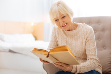 Exciting book. Pleasant cute cheerful pensioner spending a good time alone while sitting in a comfortable cozy armchair and reading an interesting book