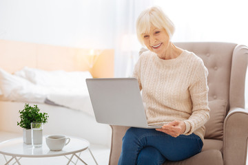 Wonderful device. Cheerful kind progressive aged woman feeling excited and happy while sitting in a cozy armchair and looking at the screen of her new modern convenient laptop