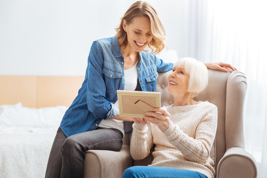 Pleasant memories. Cheerful senior woman feeling happy while sitting in a soft cozy armchair and showing a nice old photo to her kind attentive granddaughter and smiling