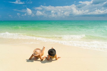 Fototapeta na wymiar Sexy bikini body of two women enjoy the sea by laying down on sand of beach wearing hat. Happy island lifestyle. White sand and crystal sea of tropical beach.
