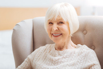 Pretty pensioner. Cute pretty senior woman feeling happy while sitting in a comfortable soft armchair and waiting for her kind young granddaughter to come and visit her