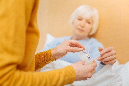 Useful pillbox. Calm serious reliable woman sitting next to her ill tired aged grandmother and giving her necessary pills from a convenient pillbox