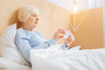 Necessary pills. Serious thoughtful ill aged woman looking concentrated while staying in bed and...
