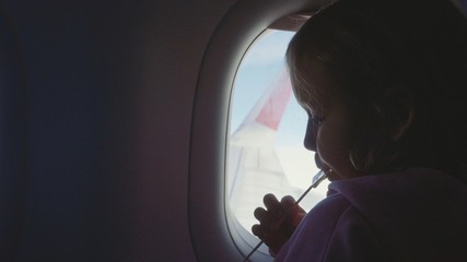 Fototapeta na wymiar Silhouette child girl sucking sweets on wooden stick over porthole in airplane