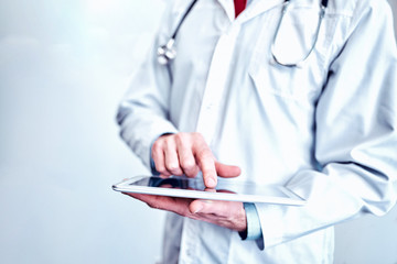 doctor in white medical coat  is using a tablet and smiling while standing against window