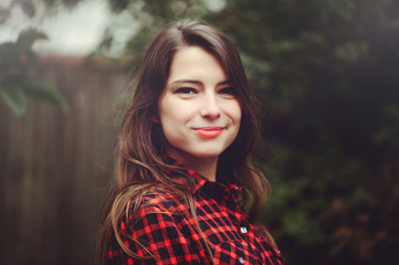 Naklejka premium Smiling young brunette woman posing in the courtyard of her residence in a red checkered shirt is happy, portrait of happy carefree, health