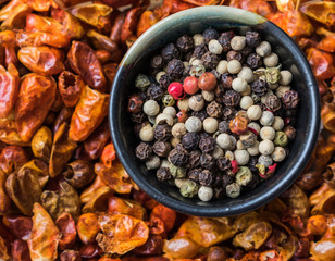 Top view of allspice, white, black, green and red pepper in a ceramic bowl over red dried chilly  peppers in the background. Spices background