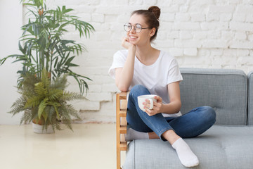 Young woman sitting on couch, chilling, drinking hot coffee and enjoying morning, takes break at...