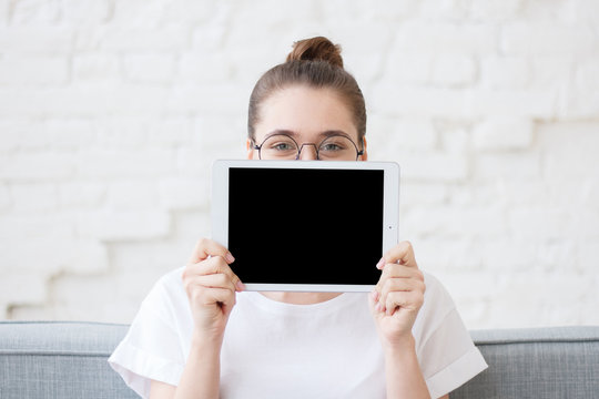 Young hipster teen with bun in round eyeglasses and white t-shirt is hiding behind digital tablet with black screen for copy space for advertising image or text