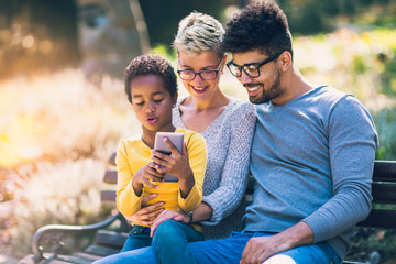Happy young mixed race couple spending time with their daughter using smart phone in public park