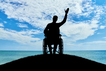 Silhouette of a disabled man in a wheelchair friendly waving his hand atop a hill near the sea