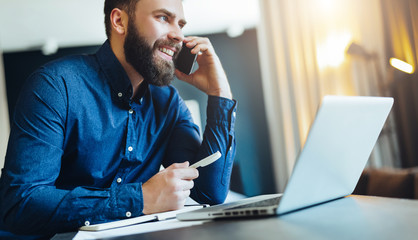 Young smiling bearded businessman is sitting at table in front of computer, talking on cell phone,...