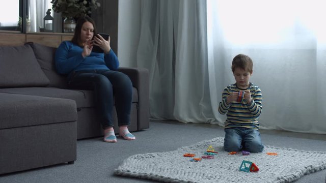 Attractive middle aged single mom browsing the net on digital tablet while cute preschool son playing with magnetic constructor toy on floor during weekend. Casual family spending free time at home.
