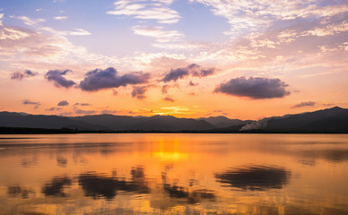 Fototapeta na wymiar Twilight scene of clouds and golden sky reflecting on flat water surface of lake surounding by dark mountain ranges.