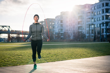 Full length of a young fit woman burning calories through alternate jumping over the skipping rope outdoors in a sunny day in the park   - obrazy, fototapety, plakaty