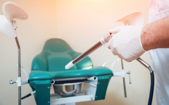 Proctologist holding an anoscope against a proctological chair.