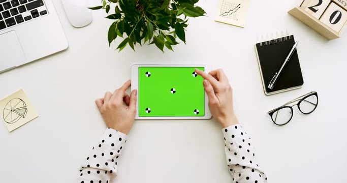 Top view on the office desk and white tablet computer with green screen and female hands taping on it. Horizontal. Office stuff beside. Chroma key. Tracking motion