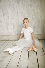 A little adorable young ballerina in white dress with point shoes sitting on woody floor and gray studio background posing on camera