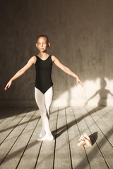 Naklejka premium A little adorable young ballerina in black sportwear and white tights exercising in the interior studio posing on camera in bright sunlight from window