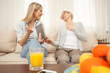 Senior mother laughing with her daughter while looking at a tablet. Happy family moments at home.