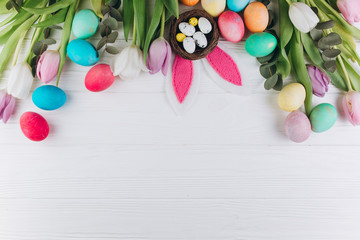 Easter composition with colored eggs, rabbit ears, nest and tulips on a white wooden background.
