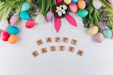 Wooden letters "Happy Easter" around colored eggs, rabbit ears, nest and tulips on a white background.