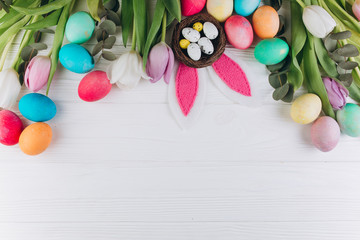 Easter composition with colored eggs, rabbit ears, nest and tulips on a white wooden background.