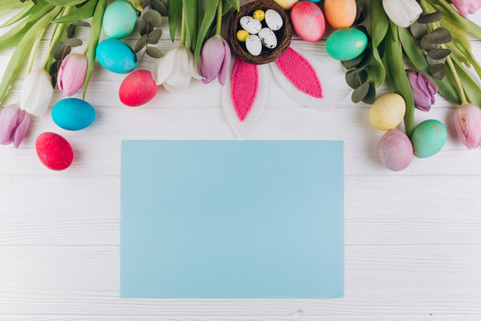 Paper with copy space around colored eggs, rabbit ears, nest and tulips on a white wooden background. Easter concept.