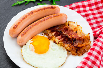 Breakfast with fried eggs, bacon, sausages and vegetable salad on dark stone background