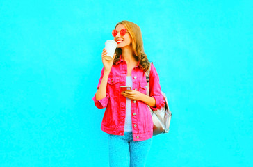 Fashion smiling woman is using smartphone in pink denim jacket holds coffee cup
