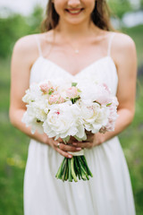 with a curly hand, the bride in a white dress, with curly flowing hair, holding in her hand a wedding bouquet of white peonies and pink roses against the background of a meadow
