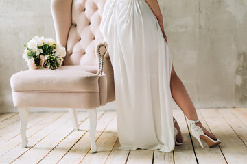 legs of the bride in white shoes and a dress that stands near the pink chair on which lies a wedding bouquet on the background of a gray wall