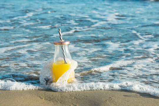 Glass Bottle with Freshly Pressed Tropical Fruits Juice with Straw Standing on Beach Sand Washed by Blue Turquoise Foamy Sea Waves. Golden Sunlight. Vacation Relaxation Freshness Summer