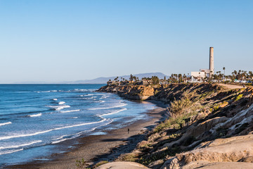 Man walks near cliffs on South Carlsbad State Beach in San Diego, California with the power plant...