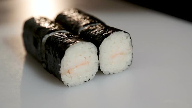 A man cuts a Japanese roll into six pieces with a knife. Roll lies on a white board. Close-up. Bare hands.