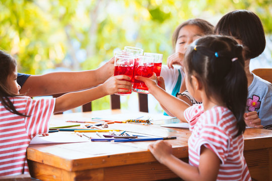 Group Of Asian Children Clinking Glasses Of Fresh Red Juice Water With Ice And Drinking Together After Finish Their Homework In The Summer Time