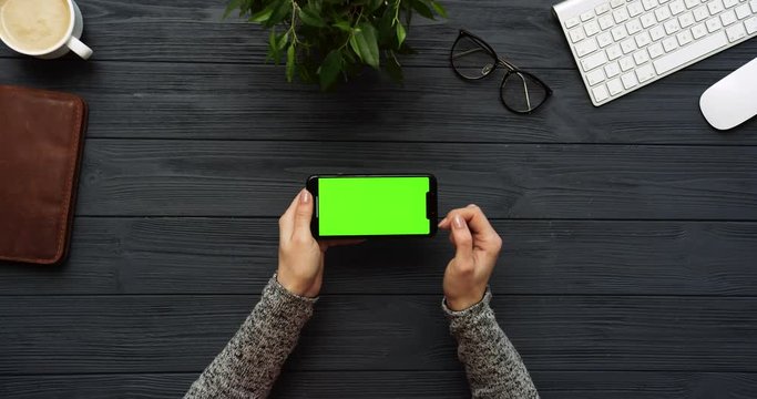 Top view on the black office desk and black smartphone with green screen and female hands taping on it. Horizontal. Office stuff beside. Chroma key.