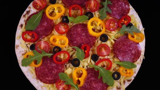 Fresh homemade pizza with salami and vegetables rotating against black background in 4K. Top view of traditional tasty food.

